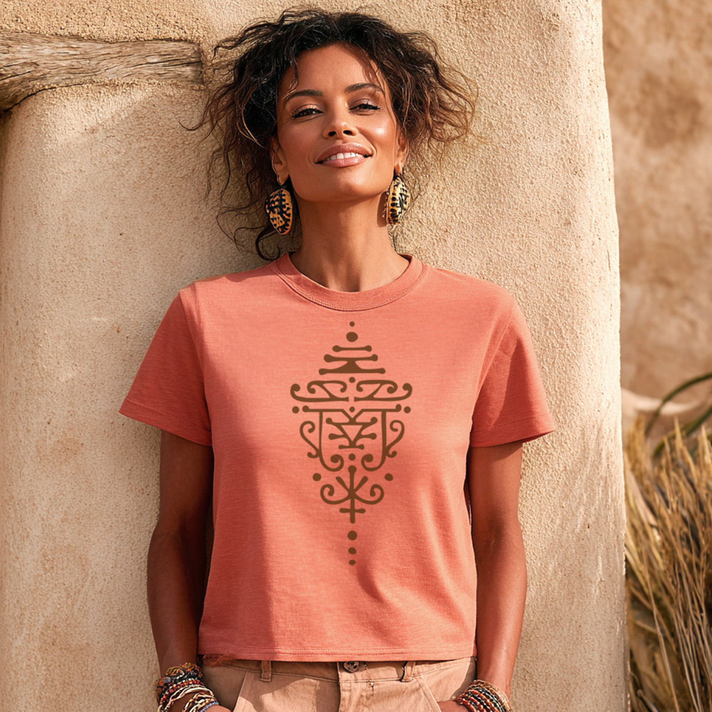 Woman wearing a terracotta t-shirt with a brown tattoo design, standing against a beige wall.